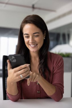 Sıraya oturmuş akıllı telefon kullanan gülümseyen ofis çalışanı, mesajlara, mesajlara, mesajlara, ya da iş ile ilgili sohbetlere cevap verir, iş başvurularından ya da sosyal medyadan gelen güncellemelere bakar