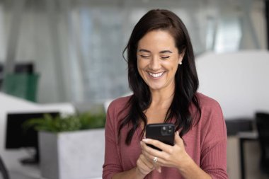 Modern ofiste akıllı telefonunu kullanırken gülümseyen, gülen, komik içerikten zevk alan, video izleyen, kaydıran ya da sohbet eden mutlu iş kadını. Yenilikçi yapay zeka iş teknolojisi kullanımı