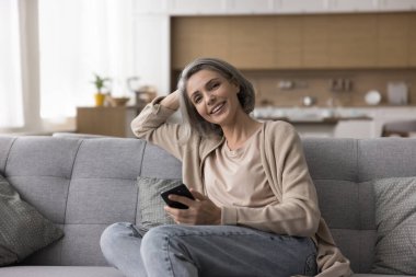 Mutlu, olgun, gri saçlı, elinde cep telefonu olan bir kadın, koltukta dinleniyor, kameraya bakıyor, içeride poz veriyor, gülümsüyor, gülüyor, dijital aletleri evdeki internet iletişimi için kullanıyor.