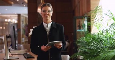 Young Hispanic woman hotel receptionist in formal suit stands at reception desk holding digital tablet, looks at camera with confident, positive expression. Customer services representative, portrait