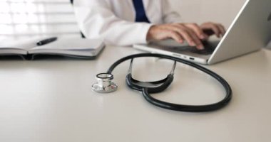 Close up of GP stethoscope on work table. Physician doctor working at computer in background, typing on laptop, writing electronic records, using telehealth service, digital technology in medicine