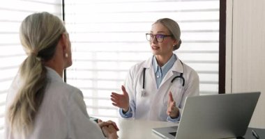 Positive young geriatric doctor in glasses meeting with female patient in hospital office, sitting at workplace table, talking to woman, giving consultation, medical advice, explaining recommendation