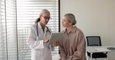 Mature grey haired doctor woman explaining medical electronic report, diagnosis, checkup result to young Caucasian patient sitting on couch, using tablet computer, giving advice