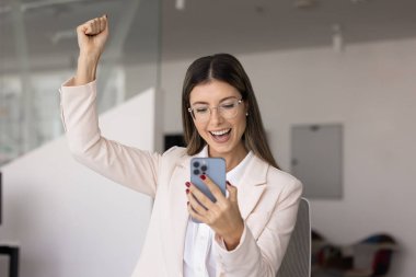 Şık gözlüklü neşeli Latin iş kadını akıllı telefondan iyi haberler alıyor, el hareketi yapıyor, cep telefonuna bakıyor, zaferi kutluyor, başarıyı kutluyor.