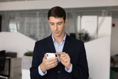 Ciddi genç Latin profesyonel bir adam cep telefonuyla yazı yazıyor, ofis yerinde aletle duruyor, sosyal medyada geziniyor, akıllı telefonda internet kullanıyor, iş servisini kullanıyor, iş için internet üzerinden iletişim kuruyor.