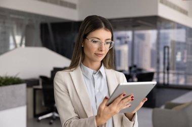 Odaklanmış genç İspanyol iş kadını gözlüklü, tablet bilgisayarına yazı yazan, ofis yerinde dijital aletlerle duran, elektronik belgeleri kontrol eden, çalışmak için online uygulama kullanan.