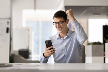 Heyecanlı gözlüklü genç Latin iş adamı zafer jesti yapıyor, cep telefonundan kazanan yumrukla mesaj okuyor, gülümsüyor, gülüyor, zaferi kutluyor, iş başarısını, geliri,