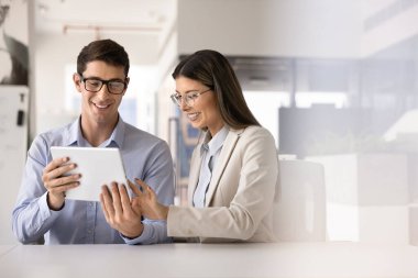 Mutlu genç yöneticiler birlikte online proje çözümü üzerinde çalışıyorlar, çalışma masasında tablet kullanıyorlar, cihaza yazıyorlar, içeriğe göz atıyorlar, gülümsüyorlar, gülüyorlar. Takım çalışması için toplanan iki meslektaş.