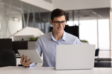 İş yerinde birden fazla alet kullanan, tablet tutan, dizüstü bilgisayarda yazan, iş yazılımıyla online ürün üzerinde çalışan, bulut çözeltisi kullanan genç, pozitif Latin bir adam.