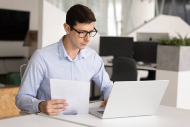 Gözlüklü genç Latin profesyonel adam bilgisayardaki muhasebe uygulamasını kullanıyor, yasal mali kağıt dökümanlarını kontrol ediyor, kağıt faturalarını ödüyor, anlaşma metninde uzmanlık yapıyor.