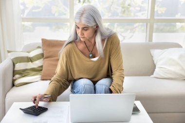 Odaklanmış olgun İspanyol ev sahibi kadın aylık giderleri hesaplıyor, iç bütçe, tasarruf, rezerv fonu, finans yönetimi için hesap makinesi ve dizüstü bilgisayarda yazma