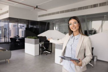 Gemiye hoş geldin diyen mutlu HR çalışanı kadın modern ofisteki boş işyerine el kaldırıyor kameraya bakıyor, gülümsüyor. Şirket işe alım adayları, yeni çalışanlar, stajyerler.