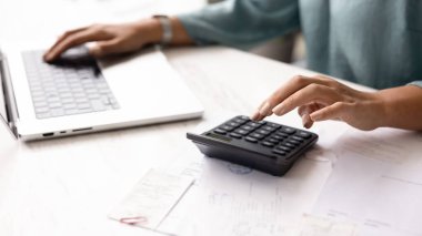 Ofis masasında hesap makinesi ve dizüstü bilgisayarda yazan kadın muhasebecinin elleri, mali plan üzerinde çalışıyor, bütçe sayımı, şirket giderleri, vergiler, ödeme faturalarını kontrol ediyor.