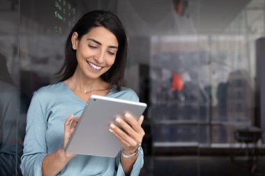 Pozitif genç Latin koç kadın ofis cam tahtasında tablet kullanıyor, cihaz ekranında daktilo yazıyor, seminer, atölye hazırlıyor, web servisi üzerinde çalışıyor, iş içi başvuru yapıyor.