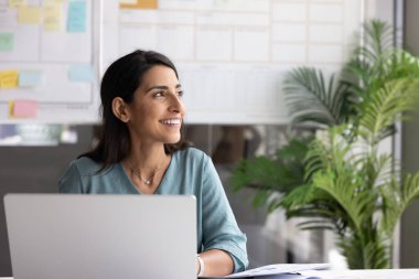 Başarılı iş fikirleri üzerine düşünen mutlu, genç İspanyol profesyonel kadın, proje için yaratıcı çözüm, dizüstü bilgisayar başında çalışmak, ofis masasında oturmak, rüya gibi dişlek gülümsemeyle başka yere bakmak.