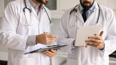 İki doktor beyaz laboratuvar önlüğü giyip hasta durumunu tartışıyor, dijital tablet kullanarak tıbbi kayıt yönetimiyle meşgul oluyor. Pratisyen hekimler, modern sağlık hizmetleri ve teknoloji arasında etkili iletişim