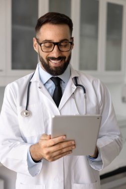 Paltoyla gülümseyen Latin kardiyolog iş yerinde tablet üzerinde çalışıyor, hasta tıbbi kayıtlarını, test sonuçlarını veya laboratuvar raporlarını inceliyor, verileri güncelliyor, reçete yazıyor, teletıp platformuna erişiyor