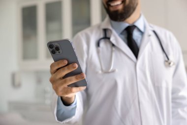 Kesilmiş beyaz önlüklü erkek doktorun görüntüsü modern akıllı telefonunu klinikte tutuyor, gülümsüyor, mesajları paylaşıyor, hasta danışmanlığı için tıbbi araçların kullanımından keyif alıyor, gerçek zamanlı destek alıyor.