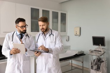 Modern muayene odasında üniforma giymiş iki çok ırklı pratisyen hekim doktor ellerinde dosya, tıp dergisi ve dijital tablet tutarak teşhis veya tedavi planı tartışmasına giriştiler. Takım çalışması.