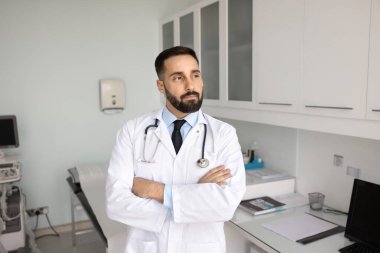 Yakışıklı Latin erkek terapist çağdaş tıbbi muayene odasında tek başına duruyor, ciddi, düşünceli bir ifadeyle başka yere bakıyor, tedavi planı üzerinde kafa yoruyor, hasta durumunu veya teşhisini dikkate alıyor.