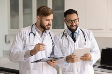 İki sağlık görevlisi ellerinde pano ve dijital tablet tutan beyaz laboratuvar önlükleri giyiyorlar, hastanede işbirliği yapıyorlar, hastaları inceliyorlar, hasta bilgilerini, tedavi planlarını veya tıbbi verileri tartışıyorlar.