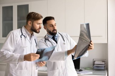 Beyaz laboratuvar önlüklü iki doktor, röntgen görüntülerini birlikte inceleyip karşılaştırıyorlar. Radyografik filmler tutuyorlar. Detaylı analiz yapıyorlar.