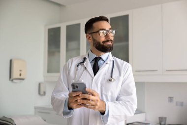 Kendine güvenen erkek doktor akıllı telefonu modern tıp ofisinde tek başına tutuyor, mobil uygulama vasıtasıyla hastayla iletişim kuruyor, dijital sağlık kayıtlarına erişiyor. Sağlık teknolojileri kullanımı