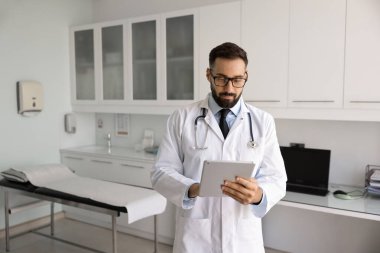 Beyaz laboratuvar önlüklü profesyonel erkek doktor dijital tablet üzerinde çalışıyor, yenilikçi dijital sağlık araçları kullanarak uzaktan iletişim kuruyor. Modern tanı ve hasta bakımında teknolojinin entegrasyonu