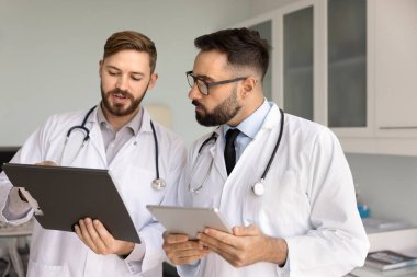 Laboratuvar önlüklü iki genç erkek doktor ellerinde dosya ve tablet tutuyorlar. İşle ilgili tartışmalar yürütüyorlar. Verimli ve etkili bir takım çalışmasıyla ilgili tıbbi bilgileri gözden geçiriyorlar.