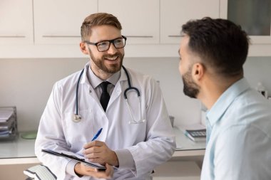 Gözlüklü ve beyaz laboratuvar önlüklü erkek pratisyen, elinde pano olan, klinikteki hastalarla istişare sırasında notlar alan, mesleki yardım, tavsiye ya da tavsiyelerde bulunan