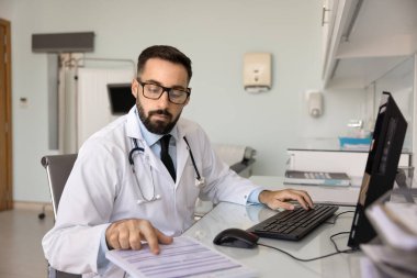 Delikanlı pratisyen hekim hasta kayıtlarını ya da tıp dergisini dikkatle inceliyor modern klinik muayene odasında bilgisayarla oturmuş, araştırma yapıyor, rapor hazırlıyor, odaklanmış görünüyor.