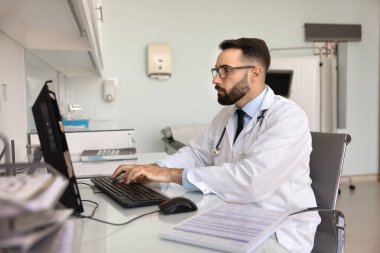 Modern tıp bürosunun masasında çalışan, elektronik sağlık kayıtlarına erişen, monitördeki bilgileri inceleyen, önemli klinik veya idari görevlere katılan, beyaz önlüklü ciddi bir doktor.