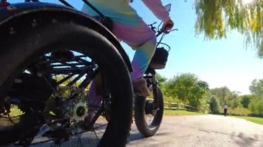 Woman in colourful candy rainbow bright unicorn pyjama or carnival dress riding an electric bicycle in the green park trail and having fun. Shiny sunny warm summertime day. Ecology concept.
