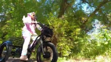 Woman in colourful candy rainbow bright unicorn pyjama or carnival dress riding an electric bicycle in the green park trail and having fun. Shiny sunny warm summertime day. Ecology concept.