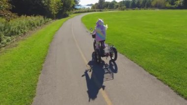 Women dressed as cartoon character unicorn on electric bicycle riding in the green sunny park. Rainbow colour silly pyjama costume. Humour and funny silly activity on the bike.