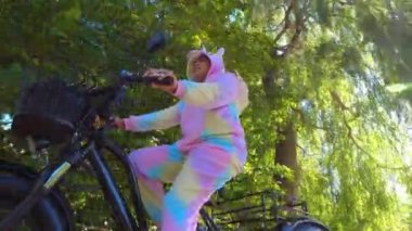 Cyclist in unicorn costume riding an electric bicycle in the green urban city area park trail. Ecology concept. Shot from the side.