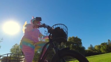 Woman in funny and crazy colourful unicorn dress riding an electric bicycle and smiling happy at sunny day. Cheerful happy woman having fun while on the e bike at green park trail. Ecology concept.