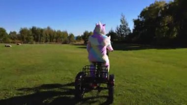 Women dressed as cartoon character unicorn on electric bicycle posing in the green sunny park. Rainbow colour silly pyjama costume. Humour and funny silly activity on the bike.