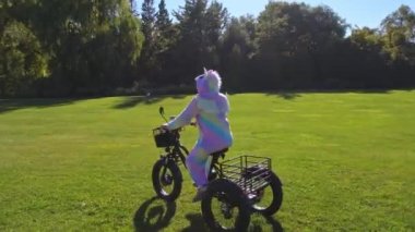 Woman in colourful candy rainbow bright unicorn pyjama or carnival dress posing on electric bicycle in the green park trail and having fun. Shiny sunny warm summertime day. Ecology concept.