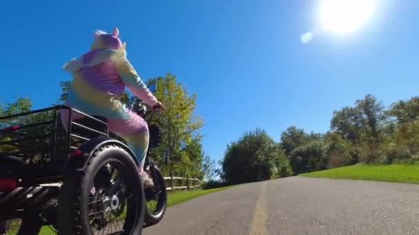 晴れた夏の日に自転車に乗るユニコーンの服を着た女性 電動自転車で魔法のユニコーンと虹色のピジャマの女の子 キュートで面白いユニコーンの服を着た人が楽しいために電子自転車に乗って — ストック動画