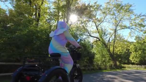 緑の日当たりの良い公園に乗って電動自転車に乗って漫画のキャラクターユニコーンとして身に着けている女性 虹色の愚かなピジャマの衣装 バイクでユーモアと面白い愚かな活動 — ストック動画