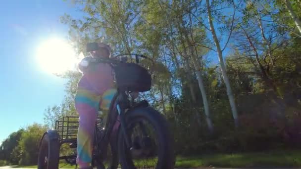 緑豊かな都市部の公園歩道で電動自転車に乗るユニコーンの衣装でサイクリスト 生態学の概念 横から撃たれた — ストック動画