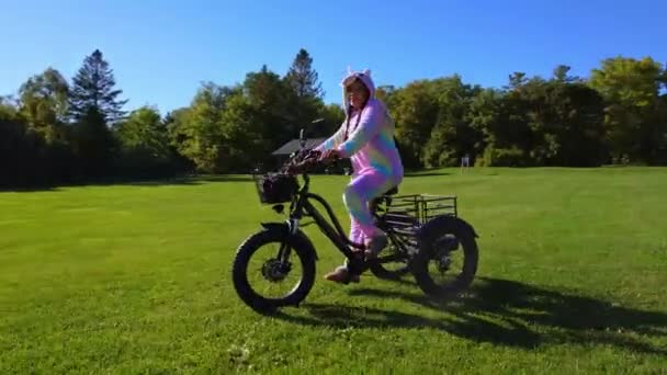 緑豊かな都市公園歩道の電動自転車に座ってユニコーンの衣装でサイクリスト 生態学の概念 横から撃たれた — ストック動画