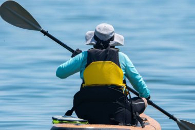 Woman paddle on board at calm lake water during warm summer. Stand up paddle board with special seating chair, wet suit and life jacket for paddling. Paddle SUP board sport and active recreation.