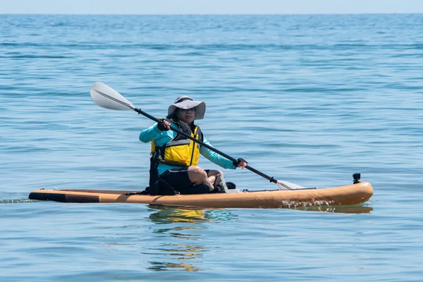 Woman sail on a SUP boards with special seating chair and wet suit and life jacket for paddling. Stand up paddle boarding active recreation leisure and sport at lake water. Active vacation.