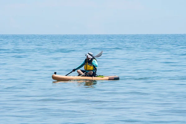 Woman paddle on board at calm lake water during warm summer. Stand up paddle board with special seating chair, wet suit and life jacket for paddling. Paddle SUP board sport and active recreation.