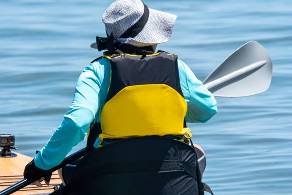 Woman paddle on board at calm lake water during warm summer. Stand up paddle board with special seating chair, wet suit and life jacket for paddling. Paddle SUP board sport and active recreation.