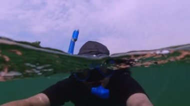 Slow motion, man swimming underwater and wearing professional wetsuit with underwater mask. Selfie or self-portrait split view while freediving in the ocean. Water and sky split view snorkelling.