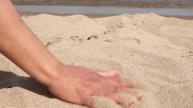 Young traveler on a sunny beach in exotic island gently stroking hot sand between her fingertips. Playful woman hand playing with yellow marine sand. Travel, adventure and leisure.