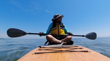 SUP 'de oturan ya da ayakta duran, dinlenen ve yolculuk sırasında su ve manzarayı gözlemleyen sağlıklı aktiviteler yapan ve büyük göllere seyahat eden bir kadın. Arkadan macera manzarası, küreklerden su damlaları